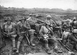 Black and white photo of six men wearing military uniform seated on a muddy slope