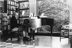 A black-and-white photograph of an elderly man with glasses sitting at a piano in front of a glass wall. Behind him is a full bookshelf.