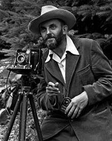 A photo of a bearded Ansel Adams with a camera on a tripod and a light meter in his hand. Adams is wearing a dark jacket and a white shirt, and the open shirt collar is spread over the lapel of his jacket. He is holding a cable release for the camera, and there is a rocky hillside behind him. The photo was taken by J. Malcolm Greany, probably in 1947.