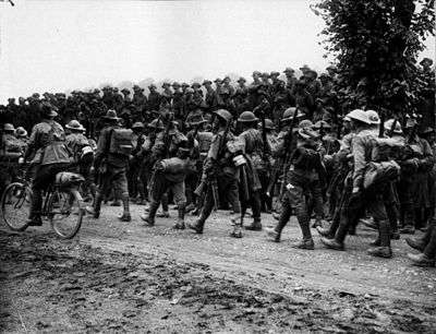 Ranks of marching men wearing Brodie Helmets and 1908 patten webbing, rifles slung being watched in the background by Australian soldiers wearing Australian slouch hats