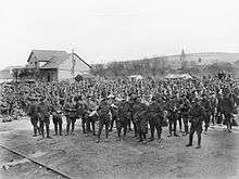 Black and white photo of a large group of men wearing military uniform standing in front of a small building. One rail of a railway track is visible in the foreground.