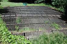 A horizontal wooden rack roughly 30 feet (9.1&nbsp;m) wide and 30 feet (9.1&nbsp;m) long lies in flat, grassy area.