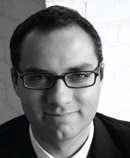 A black-and-white photograph of a man with dark hair and a cleft chin wearing glasses and looking at the viewer while smiling