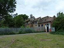Heavily overgrown brick building, with steeply sloping ground in front of it.