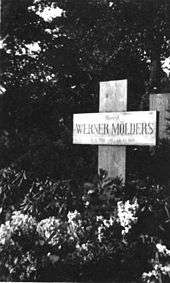 Black-and-white photograph of a wooden cross on a grave, bearing the inscription "Oberst Werner Mölders, 18. 3. 1913&nbsp;– 22. 11. 1944." The name Werner Mölders is in large letters. Trees are seen in the background; the area in front of the cross is covered with low-growing plants bearing flowers.