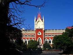 A red-and-yellow building with multiple arches and towers standing against a backdrop of blue sky and framed by trees