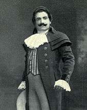 Man, with dark hair and a curling moustache, standing in a posed position. He is wearing a long coat, with lace at the throat and cuffs.