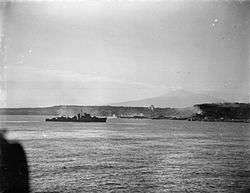 A black and white photograph showing a warship firing its armament at positions on the foreshore, while in the distance landing craft move towards the beach