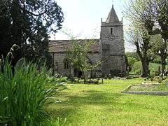 Gray stone building with small square tower and pyramidal roof. Grassy foreground with a cross and gravestones