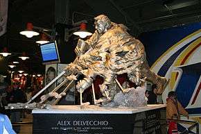 A large bronze statue of an ice hockey player in the act of shooting the puck. The player's hands and stick are shown numerous times in order to simulate motion. At the base of the statue is inscribed "Alex Delvecchio Born: December 4, 1931 Fort William, Ontario, Canada