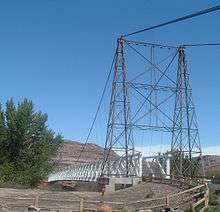 A view of a cable suspension bridge with one metal tower and a wooden deck. The second tower is partially obscured by a cottonwood tree.