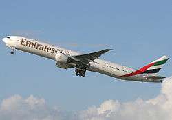 A mostly white Boeing 777, with some red, green and black markings, of Emirates, in flight, facing left.