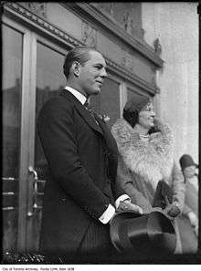 Two people standing in front of two sets of double doors, facing to the right. In front of the nearest set of doors is a man wearing a black suit with striped pants holding a top hat in front of him. In front of the far doors is an older woman wearing a fur-collared coat, clutching a purse in front of her with both hands.