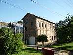Photograph of the Folsom Powerhouse, a tall, narrow masonry building with power lines overhead and the dam in the background.