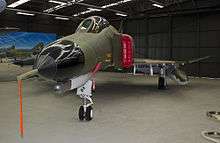 Colour photo of a military jet fighter painted in a camouflage pattern parked inside a black-walled building