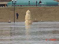 Water from a river completely surrounds a stone obelisk. In the background are several people with news cameras. A building with a green roof is seen behind them. At the bottom right of the image is the camera's time: "6 7:59&nbsp;am".