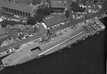 Aerial photograph taken from directly overhead of a small aircraft carrier, while she is berthed alongside a wharf. Nine aircraft are parked in a three-by-three pattern at the carrier's stern, and personnel are positioned to spell out the word "Aloha" when seen from above.
