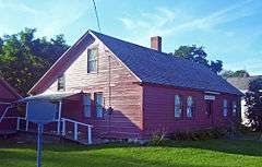 A  1&nbsp;1⁄2-story wooden house, painted red, lit by the sun from the left with trees and part of a white house behind it.