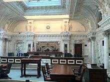 An ornate courtroom with marble columns, eleven chairs in two rows for judges, and an old fashioned wooden lectern.