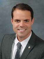 A Caucasian male with thick black hair and wearing a charcoal suit, standing against a grey background