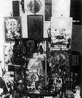 Various portraits and statuettes are arrayed on a small table and the wall it is against. A small round mirror hangs above this collection of religious items.