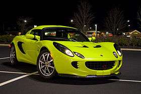 Picture of a 2005 Krypton Green Lotus Elise. The car is positioned diagonally across parking space markings in an empty parking lot of a shopping mall in Ten Broeck, Kentucky, US. A number of stores are visible in the distant background. It is night and the car is illuminated by a tall overhead lighting post to the left front of the car (not visible within the framed picture) that casts a shadow almost directly under the car, but shifted slightly to the right rear. Numerous reflected lighting highlights are visible across the smoothly curved glossy body surfaces. The photo is taken from the right front of the vehicle approximately 8-10 feet away. The front wheels are turned slightly to the left so that eight silver spokes and the center "Lotus" emblem is readily visible on the passenger side front wheel. A red vertically positioned front tow hook protrudes from the black front egg-crate grillwork and casts a slight shadow down over the front plinth. The car has been modified with the addition of a front carbon fiber splitter. The headlights and sidelights are lit.