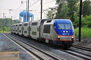 Blue and silver locomotive with orange striping pulling six bilevel locomotives on triple-track