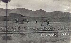 A group of men wearing sombreros walking along a dirt road between a railroad track and a line of buildings. Dated 5/13/1911.