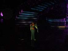 A woman is performing a song on stage. She is wearing a greenish dress.