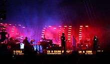 Seven musicians on a brightly lit stage sing into microphones and perform various musical instruments. They play against a multi-color background with scrolling text by their feet.