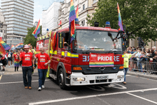 Two firefighters in red "LFB PRIDE" tshirts walk in a parade alongside a fire engine. The fire engine is decorated with at least five rainbow flags.