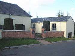 Two single-storey cream buildings with slate roofs at an angle to each other. On the left is part of the chapel; on the right is the assembly hall with two sash windows. In front is a brick wall and gateposts; above the gate is an overthrow with a lantern.