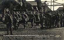 A uniformed stretcher party carry a body away from the pithead; others wait around the pithead. Wreckage from the explosion can be seen in the background.