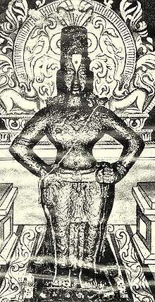 A black-and-white image of an idol of an arms-akimbo bare-chested man, wearing a conical head-gear, a dhoti and ornaments. The idol is placed on a brick, and backed by a decorated halo.