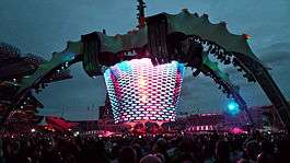 A concert stage; four large legs curve up above the stage and hold a video screen which is extended down to the band. The legs are lit up in green. The video screen has multi-coloured lights flashing on it. The audience surrounds the stage on all sides.