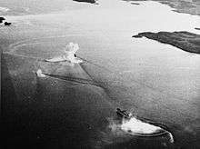 Black and white aerial photo of two ships sailing together near the shoreline. One of the ships is partially obscured by a large splash.