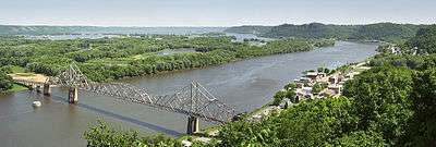 Summertime overlooking a wide and winding river. A small town is to the right. An old steel cantilever-truss bridge dominates the foreground.
