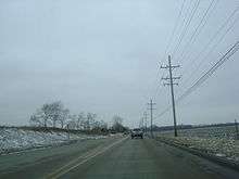 Westbound IL 120 just west of city of McHenry and narrowing to two lanes. Riding on concrete, there are open areas on both sides of the road with scattered trees on the left and utility poles on the right.