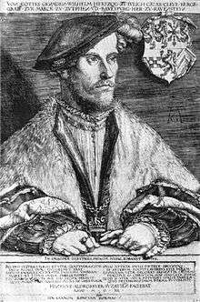 A middle aged man, Wilhelm, Duke of Jülich-Cleves-Berg, is sitting at a table. He is dressed in a soft cap that falls to the side of his head. He is wearing fur-trimmed robes, and nestled in the edges of the robes is a chain, with a cross at its nadir. His hands are folded on the table before him. He is wearing several rings, and one hand holds a pair of gloves. The family crest hangs on the chair behind him.