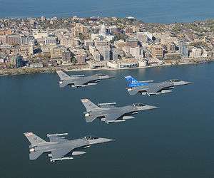 Four jets flying right in formation over water. In the foreground are buildings erected on a narrow piece of land, with water on both sides