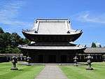Wooden building with a metal plate covered roof and an enclosing pent roof giving it the appearance of a two-storied structure.
